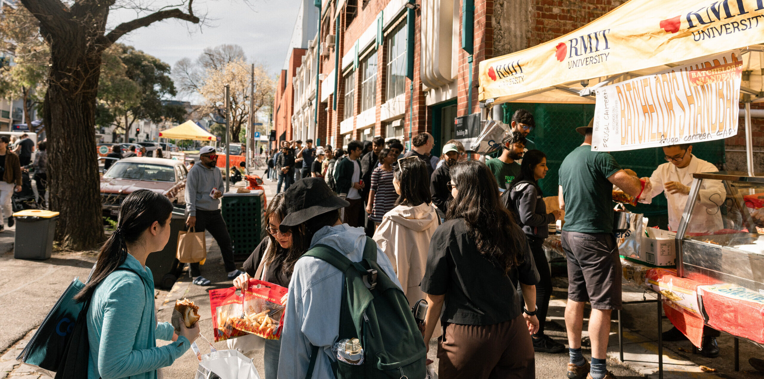A large group of people standing or walking past an RMIT stall on a sunny day.