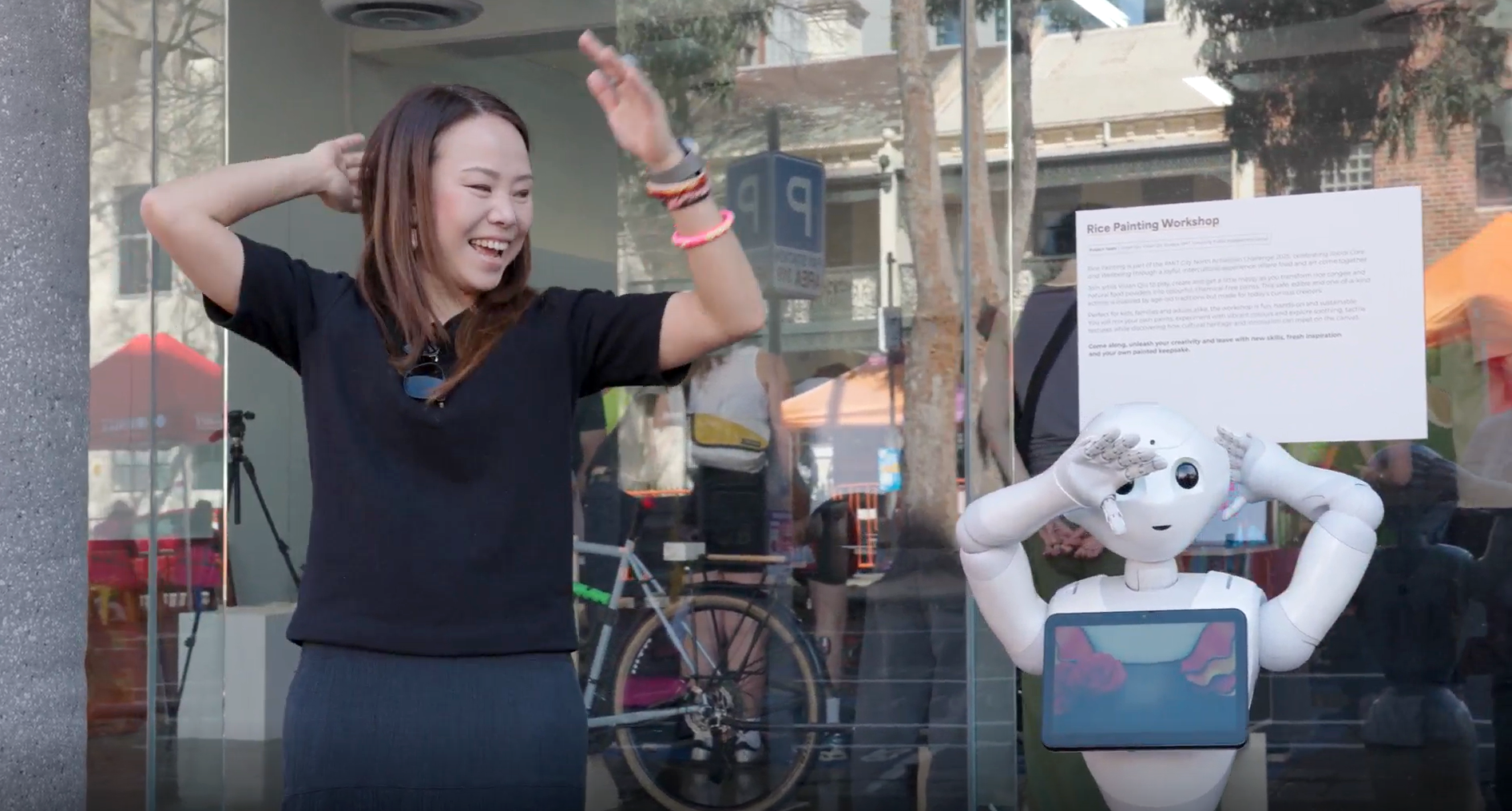 A woman smiling, standing in front of a window next to a small white robot, copying each other’s movements with their hands and arms in the air.