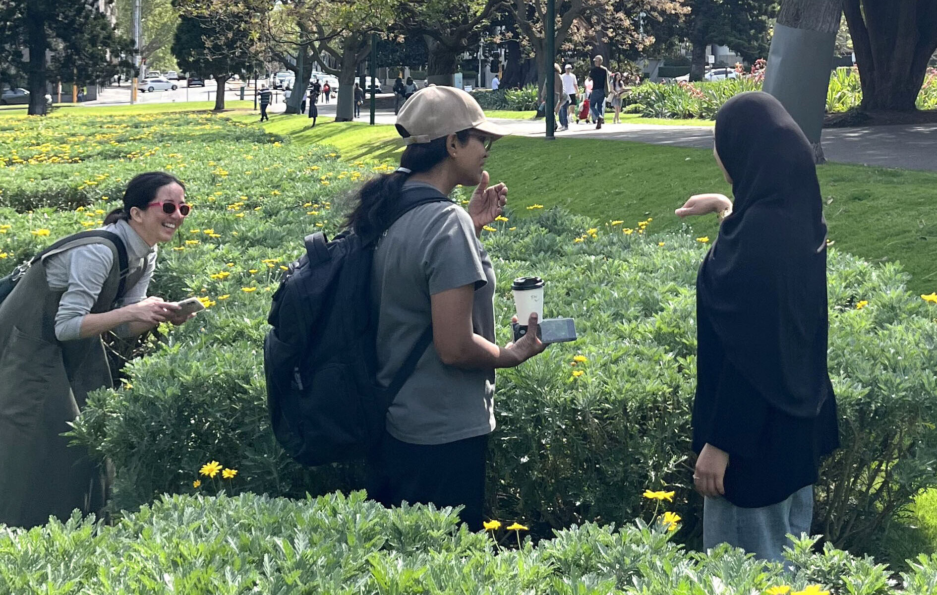 Three people are standing in a park between a large flower bed with yellow flowers. Two of the people are wearing backpacks. One is talking about the plants they are looking at.