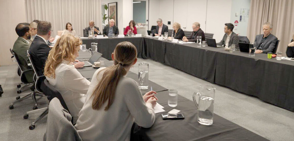 A group of people sitting around a function table with laptops, water jugs, water glasses and engaging in conversation.