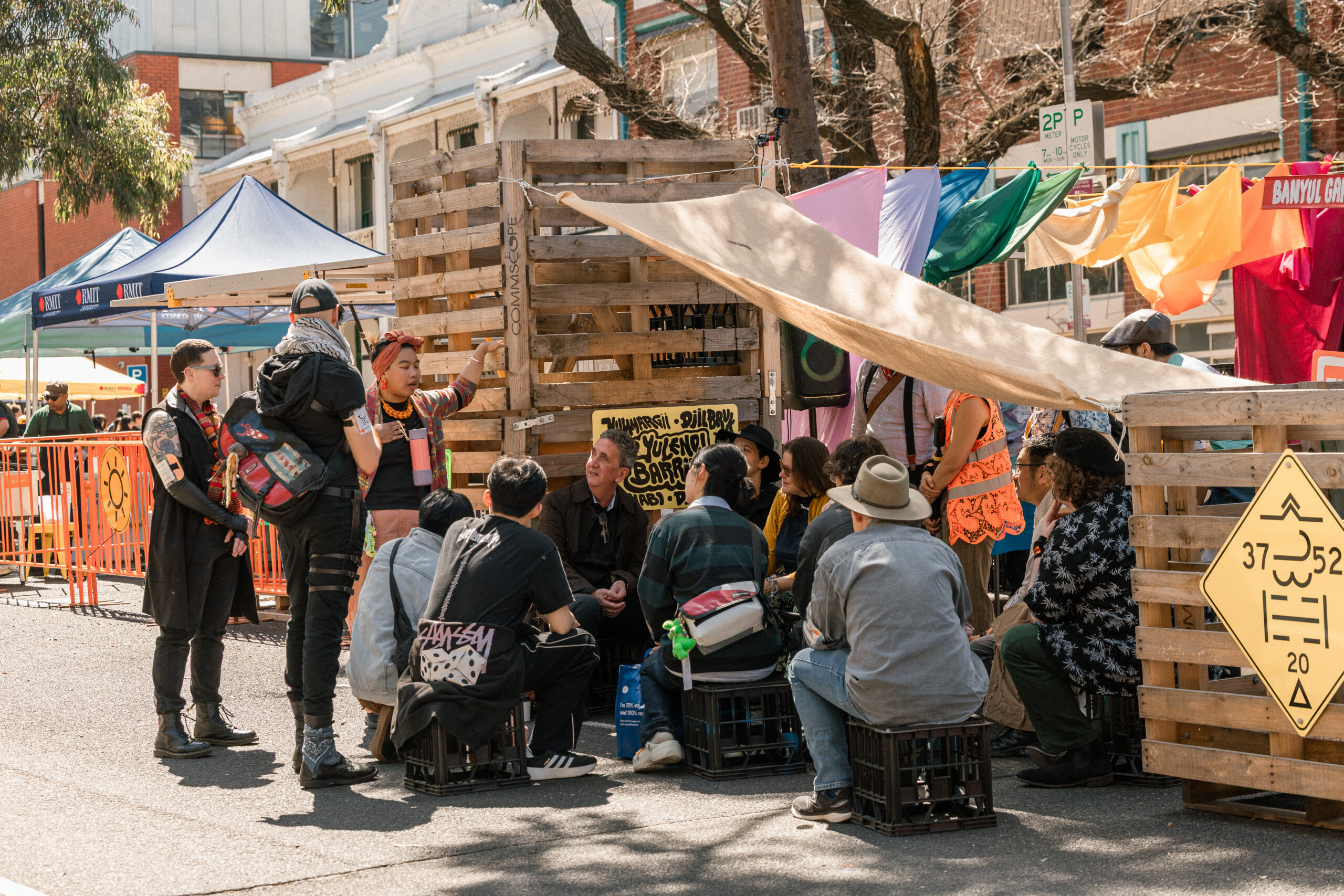 A group of people are sitting in front of a festival stall, engaging in conversation. Two people stand off to the side, chatting.